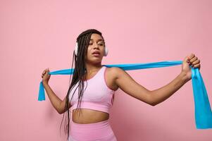 African sportswoman performs exercises with resistance band. People, sport and fitness. Young woman training with elastic strap on pink background. Active lifestyle concept photo