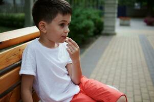 Adorable Caucasian elementary age school boy snacking outdoors, sitting on a wooden bench in the city park on summer day photo