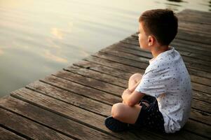 Schoolboy sitting on a pier and enjoying the tranquility of nature scene while looking at the lake landscape at sunset time photo