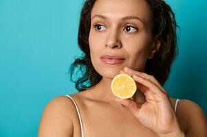 Close-up confident portrait of a natural beauty charming attractive elegant woman with curly dark hair isolated on bright blue background holding lemon half. Health care, skin treatment, body care. photo