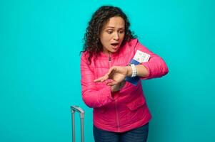 Stunned Hispanic woman in bright pink jacket looks at her watch with surprise and fear, being late for her flight, holding passport with air ticket, standing next to a suitcase against blue background photo