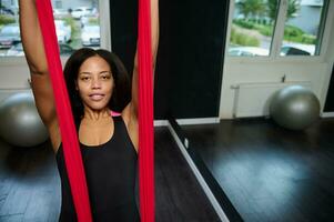 Face portrait of a young sporty slim body African woman with straps at flying aerial yoga class with red hammock photo