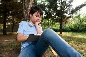 hermoso Hispano adolescente colegio chico haciendo deberes en digital tableta, sentado relajado en el parque. niños. en línea educación foto