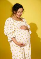 Beautiful curly haired pregnant woman in summer sundress, stroking her big belly, isolated over yellow studio background photo