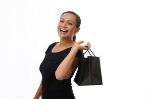 Happy excited woman dressed in black attire holds a black shopping bag and laughs, smiles with beautiful toothy smile, enjoying purchases on Black Friday, isolated on white background, copy space photo
