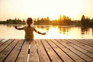 silueta de un niño sentado en el de madera muelle y disfrutando calor verano noche a el lago a puesta de sol foto