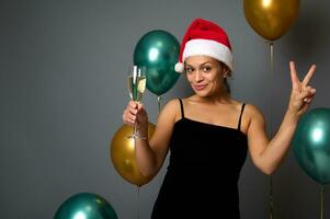 Beautiful woman in a Santa hat holds a flute with champagne and shows peace sign looking at camera against Christmas decor with luxury shiny air balloons on gray wall background with copy ad space photo