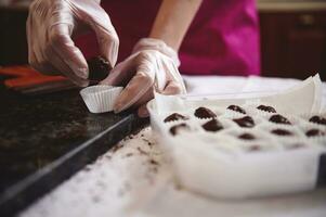 Closeup of chocolatier hands putting chocolate candy in a paper wrapper and packing chocolate truffles in box. World Chocolate Day concept. photo