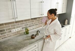 A young woman in a white waffle robe stands in the kitchen and enjoys the aroma of coffee in a jar in her hand photo