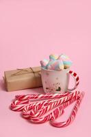 Studio shot of mug with colorful marshmallows, Christmas gift in craft wrapping paper and striped candy canes on pink colored background. Christmas and New year concept with copy space for ad photo