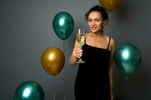 Merry Christmas and Happy New Year. Promotional shot of beautiful mixed race woman in evening dress with glass of champagne posing against gray background with shiny gold and green metallic balloons photo