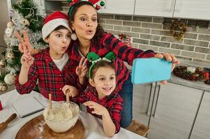 Cheerful beautiful Hispanic mom with antler hoop with her adorable children in elf hoop and Santa hat having fun together, cooking Christmas bread and making selfie looking at web camera in the phone photo