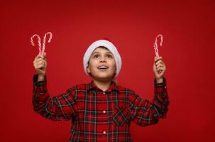 Adorable 9 years old child boy in Santa Claus hat holds sugary striped Christmas lollipops in his hands and smiles cutely with a beautiful toothy smile, looking into copy space on a red background. photo