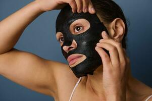 Beautiful woman puts on a hydrating smoothing nourishing cleansing black fabric mask on her face, isolated over a gray background with copy space. Skin care, cosmetology, home spa concept. Close-up photo
