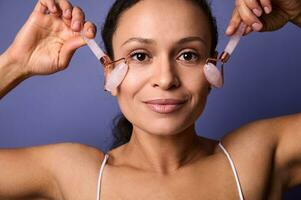 hermosa mujer con perfectamente sano limpiar piel y natural maquillaje haciendo estiramiento facial suavizado rejuvenecedor anti-envejecimiento masaje con rosado Roca rodillo masajeador aislado en púrpura antecedentes. Copiar espacio foto