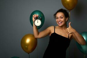 contento joven africano mujer sostiene alarma reloj, es medianoche, demostración pulgar arriba, sonriente mirando a cámara, aislado en gris antecedentes con globos nuevo año, alegre Navidad concepto para publicidad foto