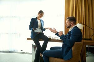 Business people negotiate in relaxed atmosphere over a cup of tea in hotel room. Successful businessman drinks coffee, listening attentively to his business partner presenting him a project on tablet photo