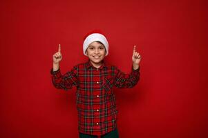 Adorable European preadolescent cheerful boy, handsome child in Santa hat and checkered shirt points with fingers up on a copy space on red colored background for Christmas and New Year advertisement photo