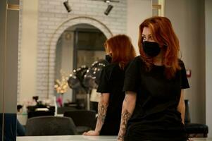 Redhead woman with tattooed arms, hair stylist wearing black uniform and safety black medical mask looking aside leaning on a countertop in front of a mirror in luxury beauty hair salon barbershop photo