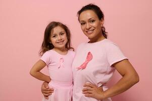 Isolated portrait on colored background with copy space of mixed race woman hugging her cute baby girl, wearing pink clothes and breast cancer awareness ribbon showing solidarity with cancer survivors photo