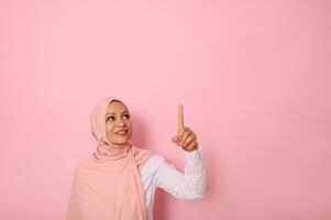 Muslim woman of Middle Eastern ethnicity dressed in religious outfit and covered head with hijab ethnicity smiles with toothy smile and looks up pointing her index finger on pink background copy space photo