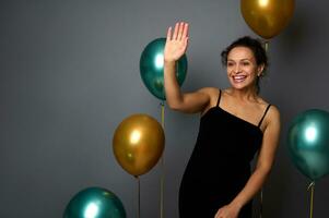 Beautiful woman dressed in elegant black evening dress stands against a gray wall background with golden and green metallic inflated balloons, waves hello with her hand, smiles with toothy smile photo