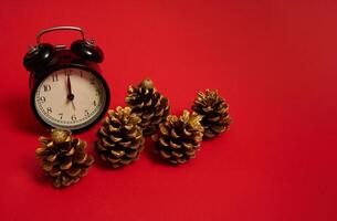 hermosa cinco pino conos decorado con oro pinturas y un negro alarma reloj con medianoche en el marcar, aislado en un de colores rojo antecedentes con Copiar espacio para anuncio. es medianoche, Navidad concepto foto