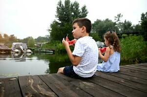 Portrait of a handsome boy eating watermelon and enjoying a beautiful summer day at sunset, sitting on the pier next to his sister, admiring the beautiful nature photo