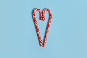 Two candy canes are laid out on a blue background in the shape of a heart. Flat lay of Christmas lollipops with copy space for ad photo
