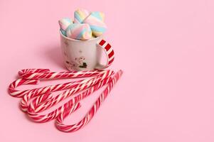 Beautiful minimalist Christmas composition with hot chocolate drink decorated with marshmallows and striped lollipops, candy canes isolated over pink colored backgroud with copy space for advertising photo