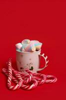 Beautiful minimalistic Christmas composition with hot chocolate drink decorated with marshmellows and striped lollipops, candy canes isolated over red colored backgroud with copy space for ad photo