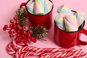 Close-up, cropped image of Christmas items, striped sugary candy canes, holly and mug with hot drink and wet colorful marshmallows on pink colored background with copy space for Christmas ad photo