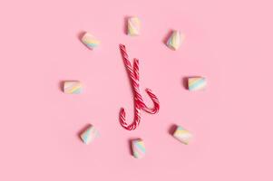 Flat lay composition with colorful sweet marshmallows and striped Christmas lollipops stacked in the shape of a clock and dial, isolated on pink pastel colored background with copy space for ad photo