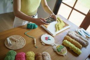 Asian Woman taking smartphone photo Punch needle. phone posting on social networks in studio workshop. designer workplace Handmade craft project DIY embroidery