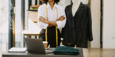 Female fashion, designer, Looking at Drawings and Sketches that are Pinned to the Wall Behind Her Desk. Studio is Sunny. Personal Computer, Colorful Fabrics, Sewing Items are Visible. photo