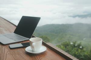 Young man freelancer traveler wearing hat anywhere working online using laptop and enjoying mountains view photo