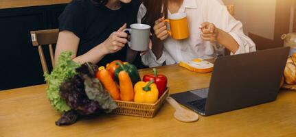 Happy two young women looking laptop computer during cooking together in kitchen room at home. Two young diverse lesbian women spending time together. LGBT and gender identity concept photo