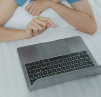 Young Asian woman with beautiful face, long hair, blue shirt lying on bed in white bedroom at home with cup of coffee and video call with laptop talking to relatives on vacation. holiday concept. photo