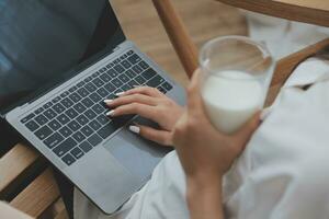 Happy young female in casual clothes smiling and looking at camera while using laptop on bed in bedroom at home photo