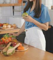 A young woman with a beautiful face in a blue shirt with long hair eating fruit sitting inside the kitchen at home with a laptop and notebook for relaxation, Concept Vacation. photo