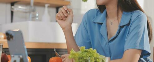 A young woman with a beautiful face in a blue shirt with long hair eating fruit sitting inside the kitchen at home with a laptop and notebook for relaxation, Concept Vacation. photo