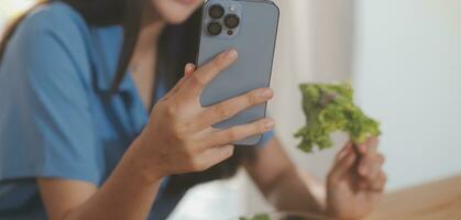 un joven mujer con un hermosa cara en un azul camisa con largo pelo comiendo Fruta sentado dentro el cocina a hogar con un ordenador portátil y cuaderno para relajación, concepto vacaciones. foto