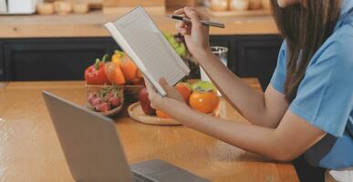 A young woman with a beautiful face in a blue shirt with long hair eating fruit sitting inside the kitchen at home with a laptop and notebook for relaxation, Concept Vacation. photo