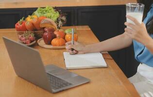 A young woman with a beautiful face in a blue shirt with long hair eating fruit sitting inside the kitchen at home with a laptop and notebook for relaxation, Concept Vacation. photo