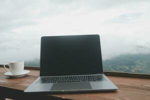 computadora monitor, teclado, café taza y ratón con blanco es en el trabajo mesa a el cielo montaña río y arboles frente ver antecedentes. foto