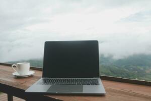 computadora monitor, teclado, café taza y ratón con blanco es en el trabajo mesa a el cielo montaña río y arboles frente ver antecedentes. foto