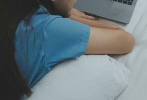 Young Asian woman with beautiful face, long hair, blue shirt lying on bed in white bedroom at home with cup of coffee and video call with laptop talking to relatives on vacation. holiday concept. photo