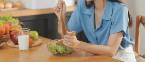 A young woman with a beautiful face in a blue shirt with long hair eating fruit sitting inside the kitchen at home with a laptop and notebook for relaxation, Concept Vacation. photo