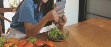 A young woman with a beautiful face in a blue shirt with long hair eating fruit sitting inside the kitchen at home with a laptop and notebook for relaxation, Concept Vacation. photo