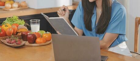 A young woman with a beautiful face in a blue shirt with long hair eating fruit sitting inside the kitchen at home with a laptop and notebook for relaxation, Concept Vacation. photo